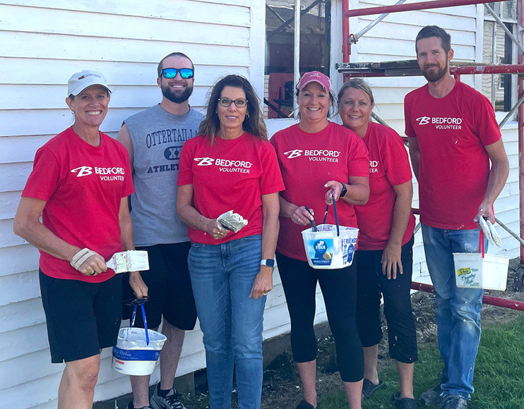 4 women and 2 men in matching red "Bedford Volunteer" T-Shirts hold paint brushes near an old building and smile at the camera. 