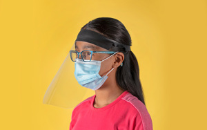 A young girl wearing glasses and a face mask models a clear face shield manufactured by Bedford Industries