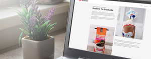 A laptop screen shows a "Bedford Tie Products" webpage with product images. A potted plant is nearby. The Bedford logo is in the corner.