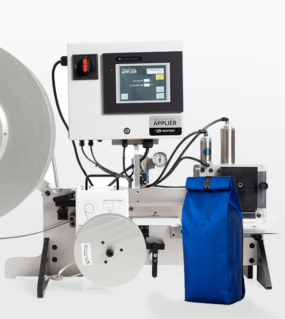 A silver table top machine that applies bag closures vertically with a blue coffee bag in front of the machine.