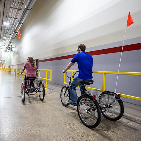A man and a woman pedal bicycles in a manufacturing facility.