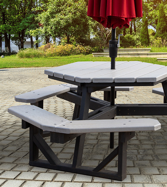 Hexagonal plastic lumber picnic table with gray top and black frame, red umbrella, on a stone patio in a park setting.