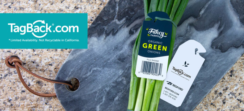 A close-up, angled shot of a bunch of organic green onions on a gray marble cutting board. The onions are tied with a dark blue tag that reads 'Fancy Farms Organic Green Onions' and includes a barcode and the PLU code '94068'. Next to the onions is a white tag with the 'TagBack.com' logo and the Bedford company information, including their website and phone numbers. In the upper left corner of the image is the 'TagBack.com' logo in a blue rectangle with the disclaimer '*Limited Availability. Not Recyclable in California.'