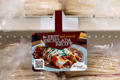 A shelf talker recipe card for "the BEST and easiest ENCHILADA RECIPE" hangs in a grocery store, with packages of tortillas visible behind it.