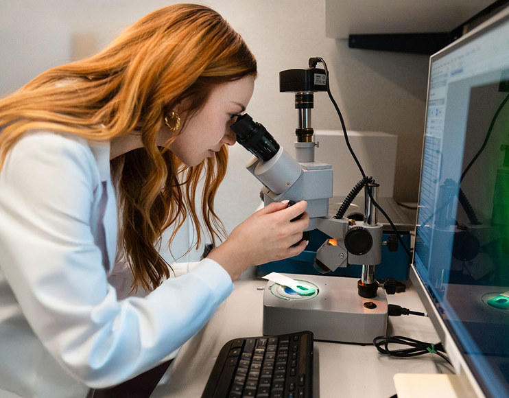 A woman in a white lab coat looks into a microscope. 