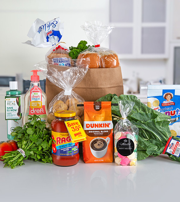 A still life photograph of various grocery items arranged on a light-colored countertop against a blurred kitchen background. The items are spread out and include: a white plastic bag with 'Soft White' branding floating in the upper left; a small paper bag containing baked goods wrapped in clear plastic, topped with a red tie; a clear plastic bag filled with dinner rolls in the center; a bottle of green liquid with 'REAL SAVE $2 NOW 100%' written on it; a bottle of 'dreft' dish soap; a jar of 'RAGU Old World Style TRADITIONAL SAUCE' with a 'Save 35¢' sticker; a bag of 'DUNKIN' ORIGINAL BLEND' coffee with a picture of a coffee cup; a clear bag of 'Abdallah MINT MELLETS' candies; a box of 'Little Debbie Powdered Mini Donuts'; a bunch of leafy greens; a red tomato; and a bunch of cilantro. The background shows a white kitchen counter with blurred cabinets and a window.