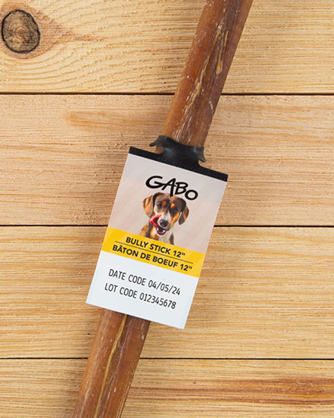 Gabo Bully Stick 12" dog treat with tag showing dog image, date/lot code. On wooden planks.