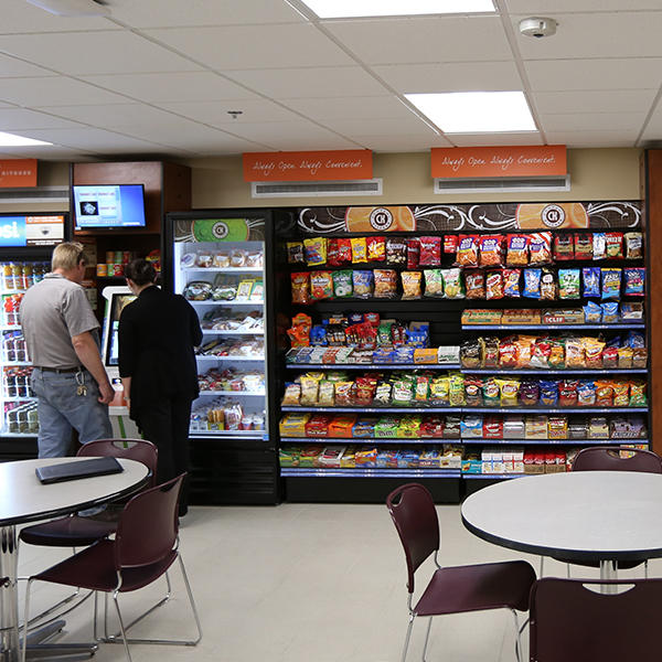 A man and a woman make food selections from a break room display of snack items.