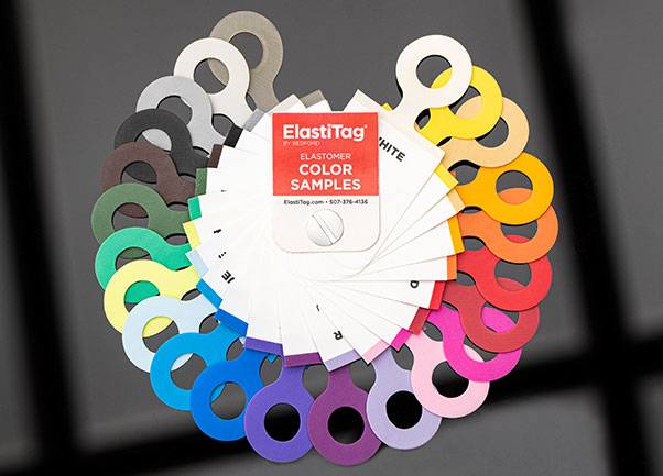 Elastomer swatch color wheel A fan-shaped display of ElastiTag color samples. The samples are arranged in a rainbow pattern, with each color represented by a small, circular tag. The tags are attached to a white card with the ElastiTag logo. The colors shown include white, yellow, orange, red, pink, purple, blue, green, and black.
